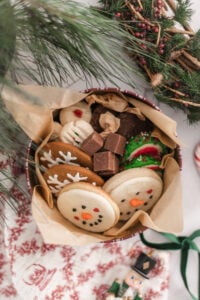A festive tin filled with decorated holiday cookies, including snowmen, snowflakes, and Christmas trees, along with pieces of chocolate, surrounded by pine branches and a holiday wreath.