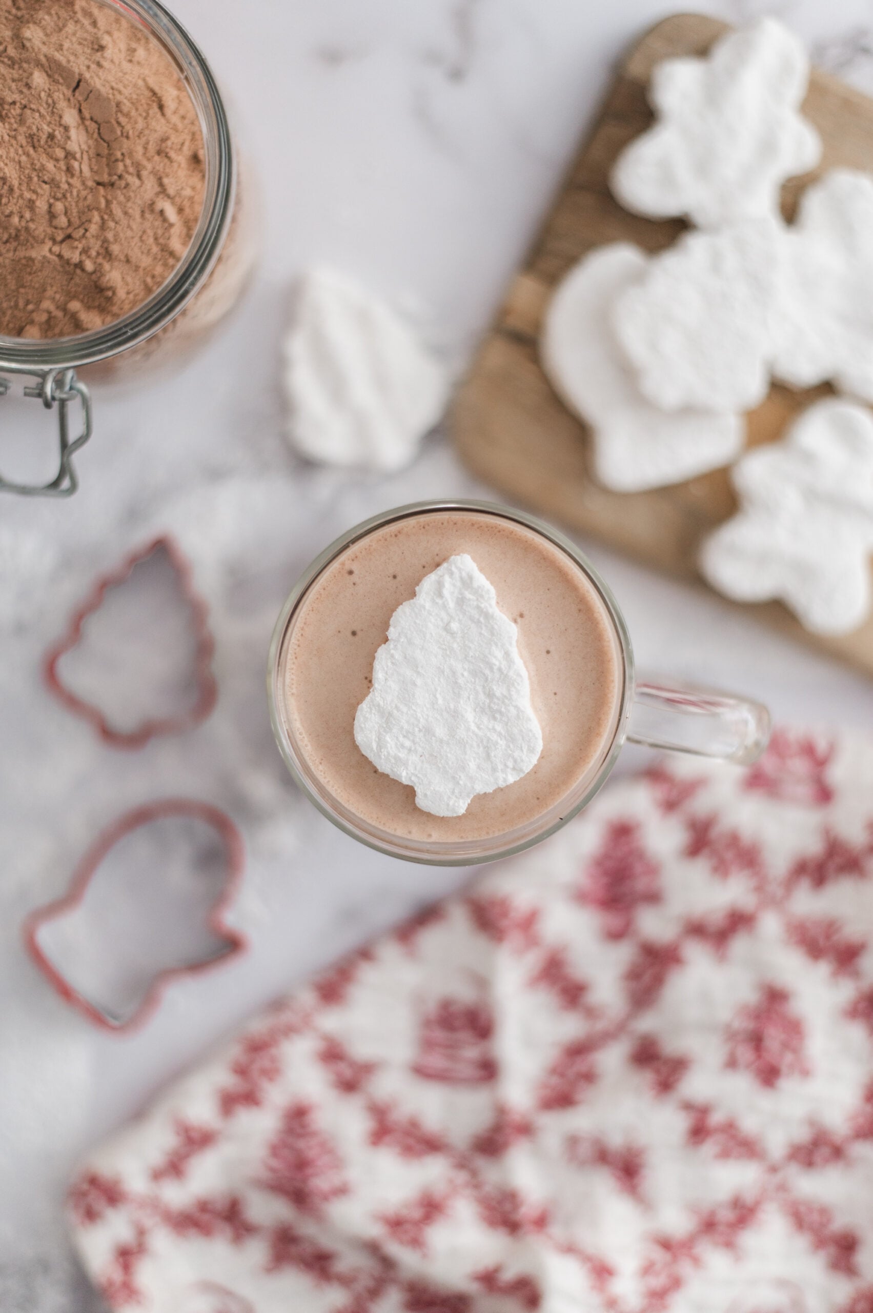 A mug of hot chocolate topped with a tree-shaped marshmallow, with more marshmallows, a jar of cocoa powder, cookie cutters, and a red patterned cloth nearby.