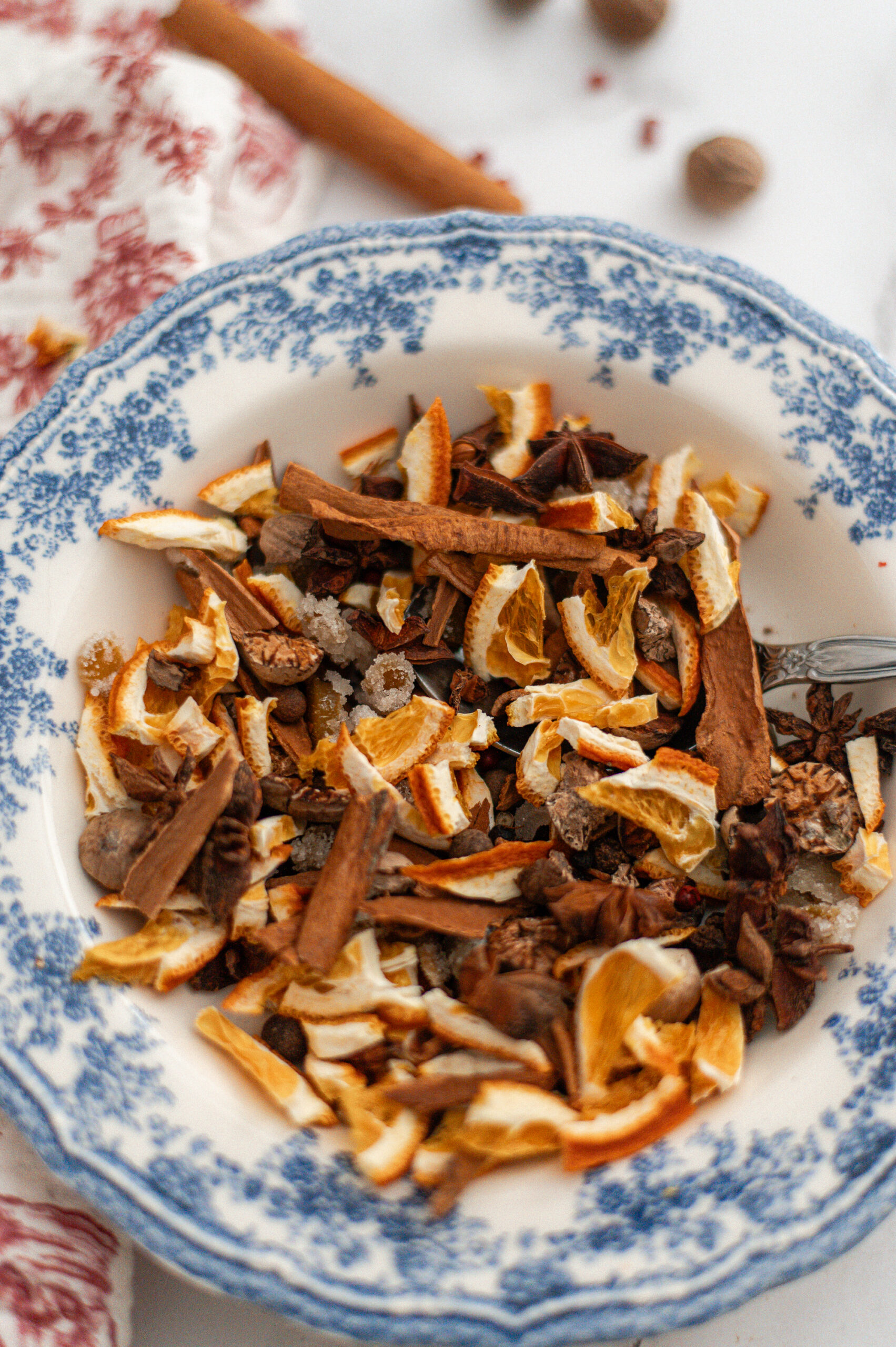 A blue and white patterned bowl filled with dried orange peels, cinnamon sticks, star anise, cloves, and other spices on a light surface. A spoon is partially visible on the right side of the bowl.