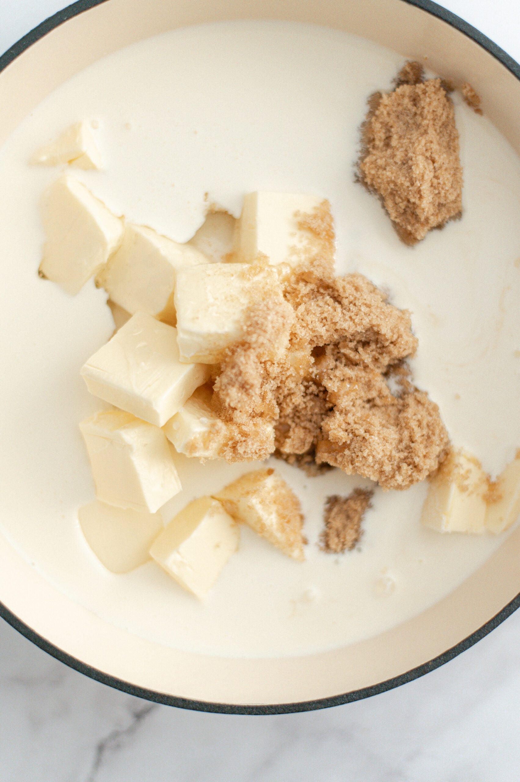 A close-up of a pot containing heavy cream, chunks of butter, and scoops of light brown sugar, ready to be mixed and cooked into delicious homemade salted caramel.