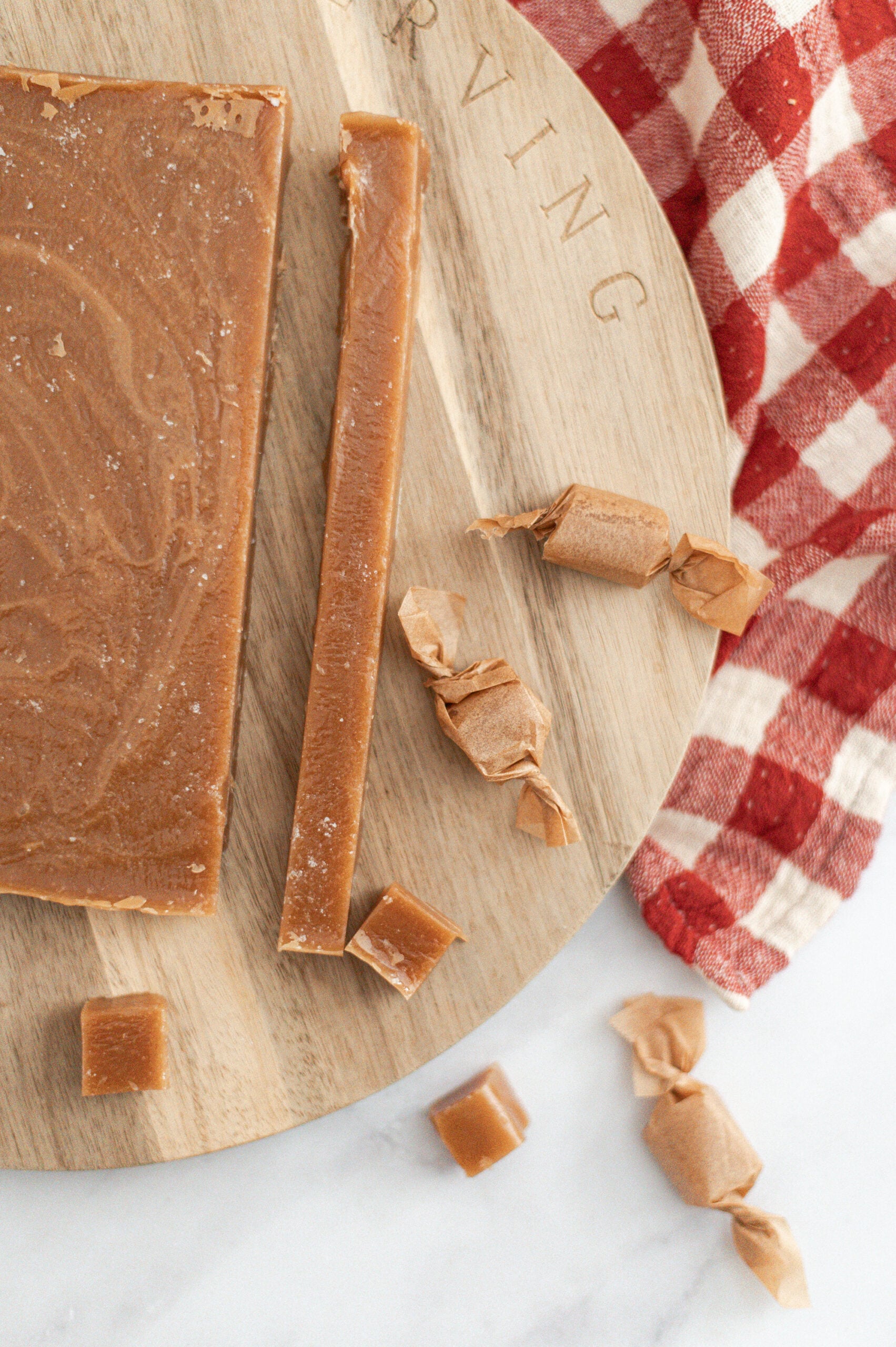A wooden board holds a slab of homemade salted caramel fudge, with one long strip sliced and several small caramel pieces, some wrapped in parchment paper, beside a red and white checkered cloth on a marble surface.