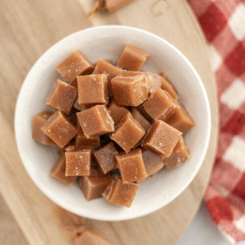 A white bowl filled with square homemade salted caramel candies sits on a wooden board, next to a red and white checkered cloth. Several wrapped caramels are scattered nearby.