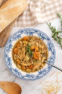 A bowl of Homestyle Chicken Noodle Soup with shredded chicken, carrots, and savory broth, garnished with rosemary. The bowl rests on marble beside a wooden spoon, bread, uncooked noodles, and a checkered cloth for a cozy touch.