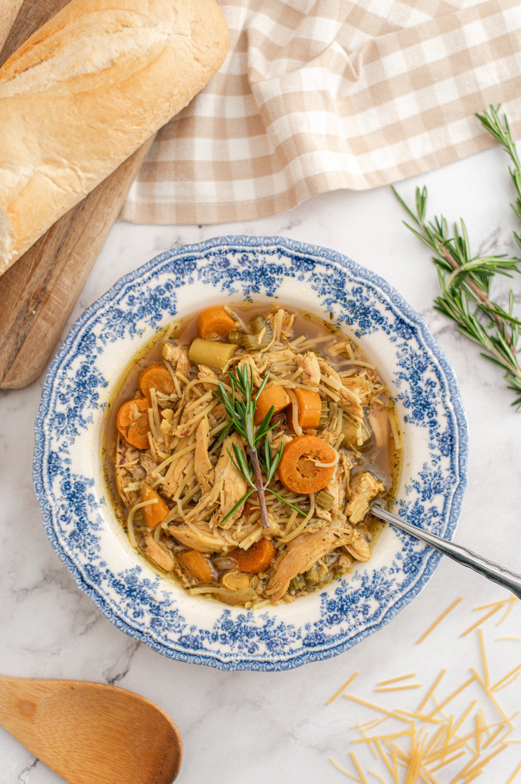 A bowl of Homestyle Chicken Noodle Soup with shredded chicken, carrots, and savory broth, garnished with rosemary. The bowl rests on marble beside a wooden spoon, bread, uncooked noodles, and a checkered cloth for a cozy touch.