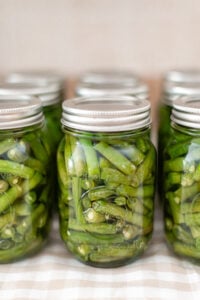 Several glass jars filled with pickled green beans are arranged in rows on a checkered cloth. The jars, sealed with metal lids, showcase the results of learning how to can green beans in a pressure canner.