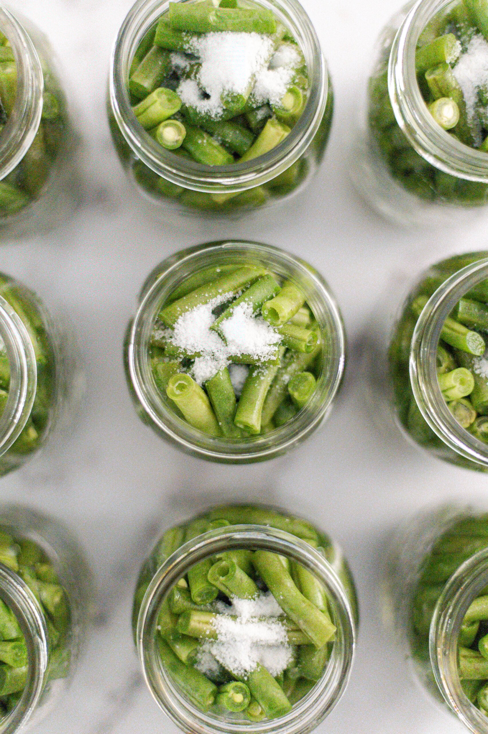 Overhead view of glass jars filled with cut green beans and topped with granules of salt, arranged in a neat grid pattern on a white surface—perfect for illustrating how to can green beans in a pressure canner.