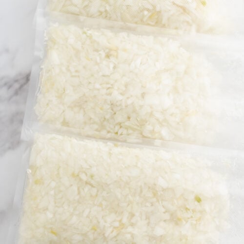 Three vacuum-sealed bags filled with chopped onions are stacked on a tray with a white marble background.