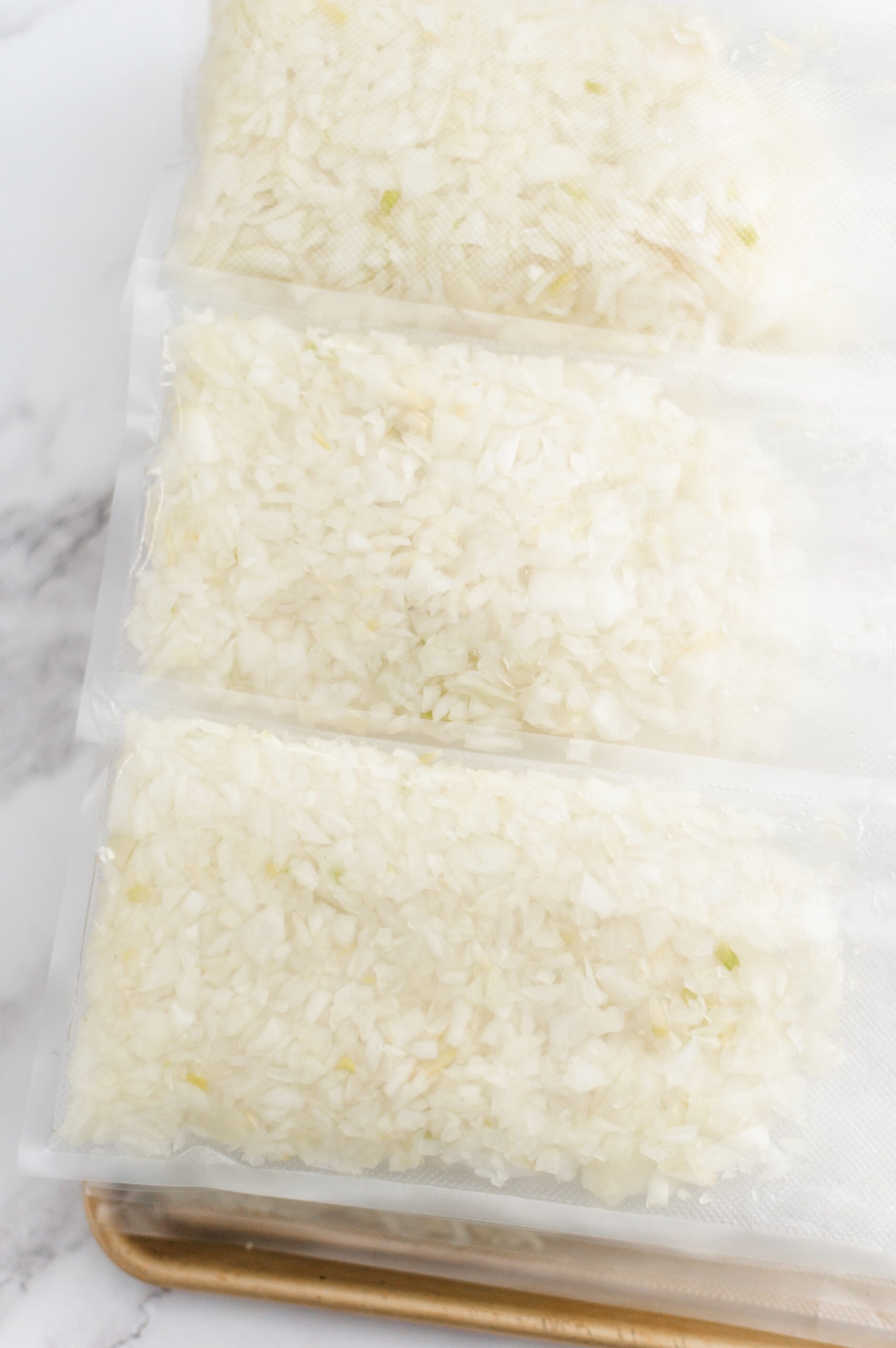 Three vacuum-sealed bags filled with chopped onions are stacked on a tray with a white marble background.