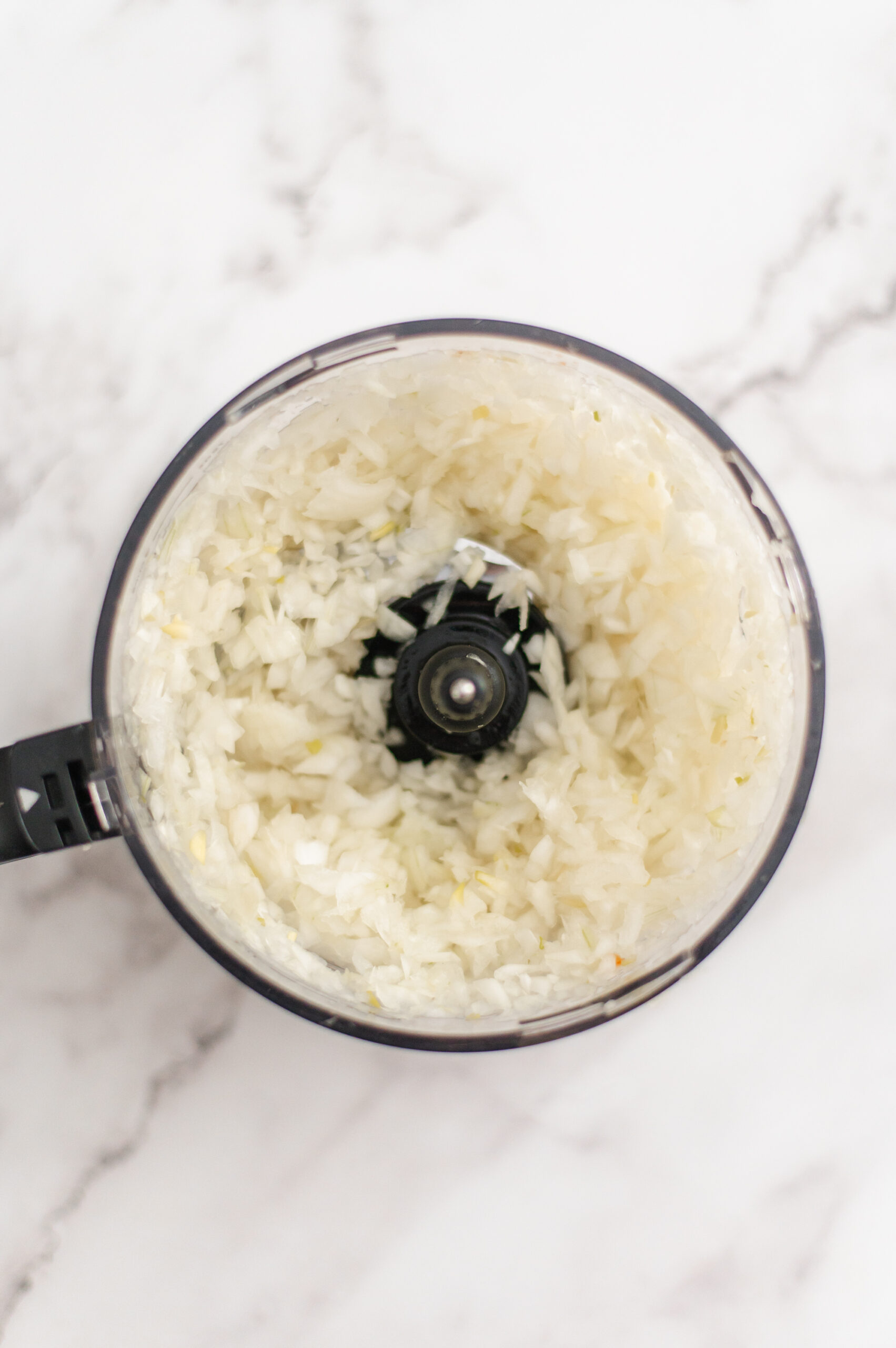A top-down view of finely chopped onions inside a food processor bowl, placed on a white marble countertop.