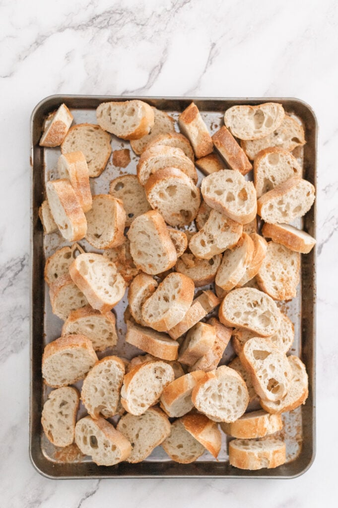 A baking sheet filled with unevenly arranged slices of baguette bread, perfect for learning how to make homemade breadcrumbs, placed on a white marble surface.