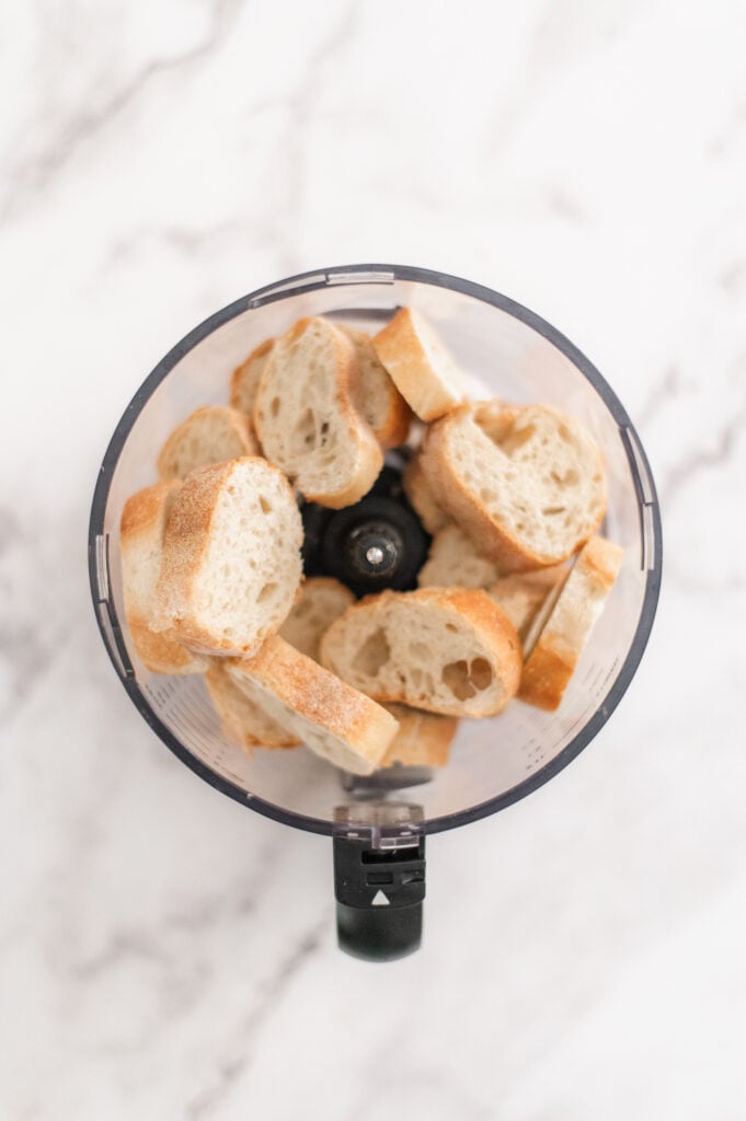 Slices of bread placed inside a food processor, viewed from above on a white marble surface—perfect for learning how to make homemade breadcrumbs.