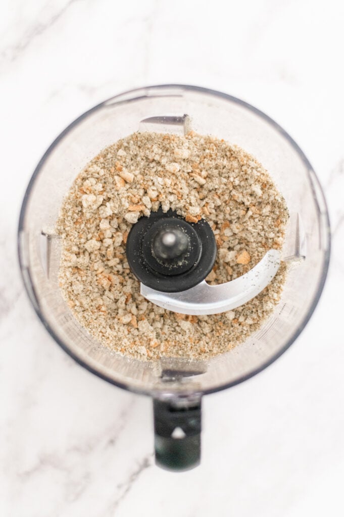 A top-down view of a food processor containing finely ground crumbs, perfect for anyone learning How to Make Homemade Breadcrumbs, sitting on a white marble surface.
