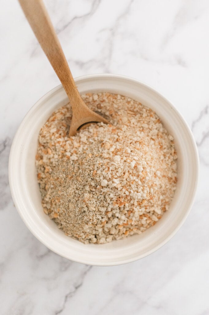 A white bowl filled with breadcrumbs sits on a marble surface, with a wooden spoon resting inside the bowl—perfect for showing How to Make Homemade Breadcrumbs.