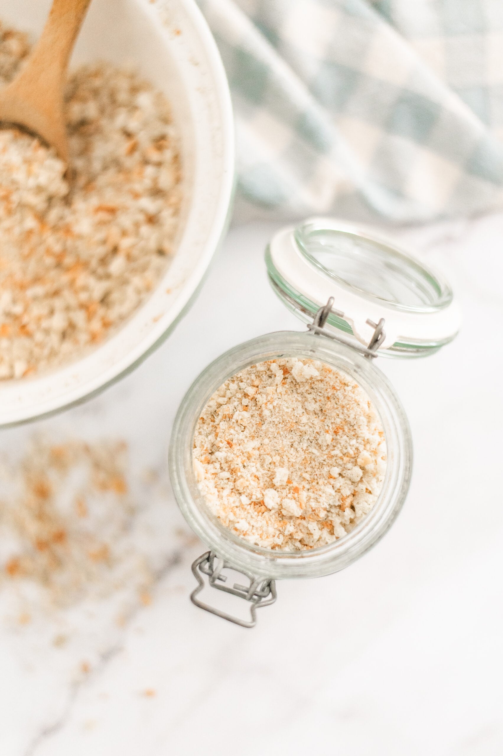 A glass jar filled with homemade breadcrumbs sits on a marble surface beside a bowl of breadcrumbs being mixed with a wooden spoon—a perfect scene for showing How to Make Homemade Breadcrumbs. A blue and white checkered cloth is in the background.