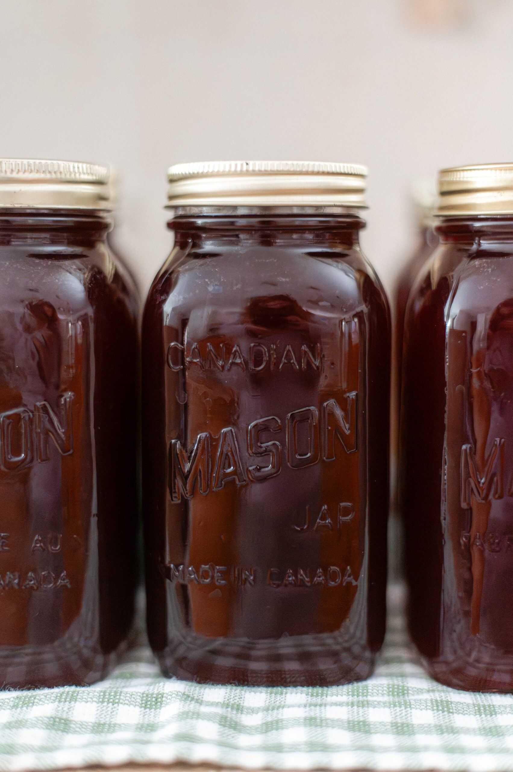 Three mason jars filled with dark brown liquid, possibly homemade grape juice, are lined up on a green and white checkered surface. The gold lids and “CANADIAN MASON” embossed on the glass hint at learning how to make and can grape juice at home.