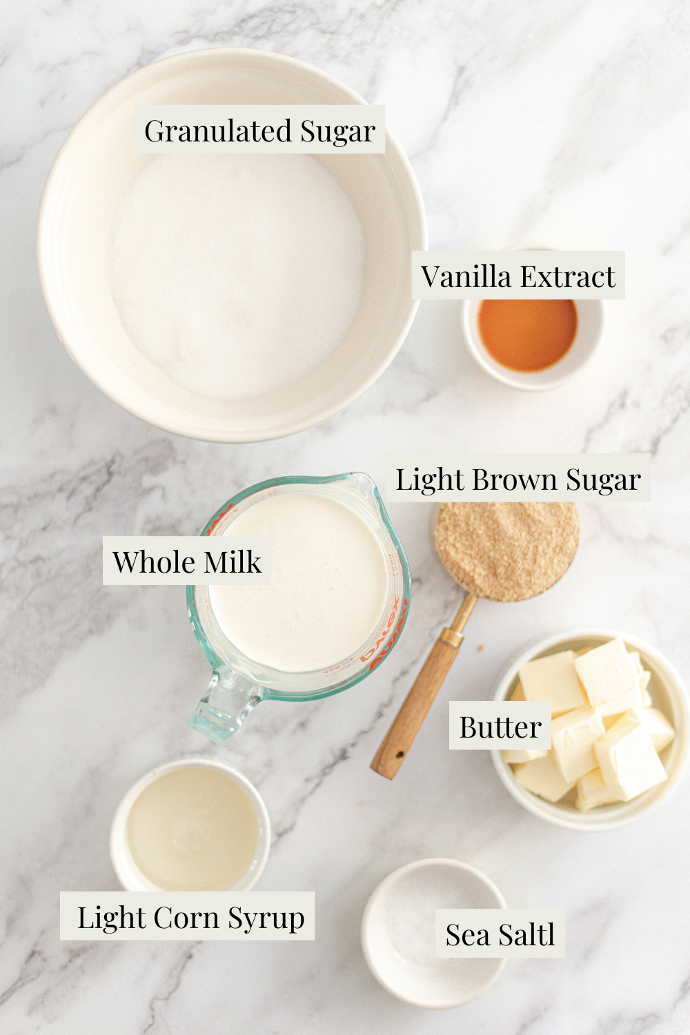 A marble surface with labeled bowls and a measuring cup holding ingredients for Homemade Salted Caramel—granulated sugar, vanilla extract, light brown sugar, whole milk, butter cubes, light corn syrup, and sea salt—neatly arranged for a recipe.