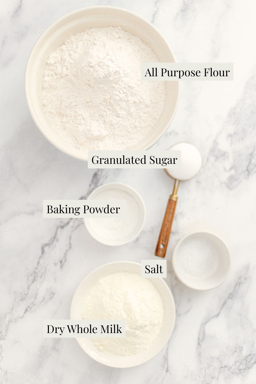 Five labeled bowls on a marble surface containing all-purpose flour, granulated sugar, baking powder, salt, and dry whole milk, with a measuring spoon next to the salt bowl.