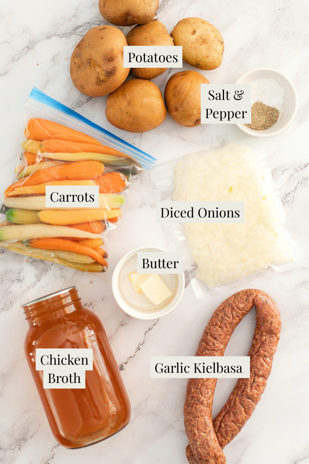 Ingredients for a recipe displayed on a white surface: potatoes, a small bowl of salt and pepper, carrots in a bag, diced onions in a bag, a dish of butter, a jar of chicken broth, and two garlic kielbasa sausages. Labels identify each item.
