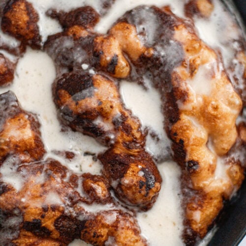 Close-up of a skillet dessert with golden-brown dough pieces covered in melted vanilla icing and swirls of cinnamon, resembling a freshly baked monkey bread or cinnamon roll casserole.