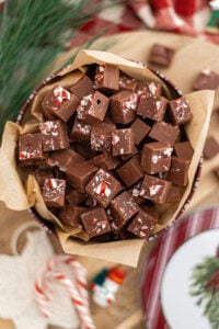 A tin lined with parchment paper is filled with squares of Simple 3 Ingredient Homemade Fudge, each topped with crushed peppermint candy. Holiday decorations and candy canes are visible in the background.