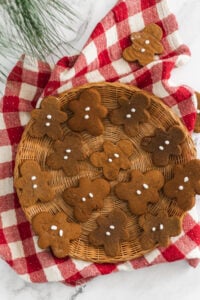 A round wicker tray filled with gingerbread cookies shaped like people, decorated with simple white icing dots, sits on a red and white checkered cloth on a white surface. Some pine needles are visible in the corner.