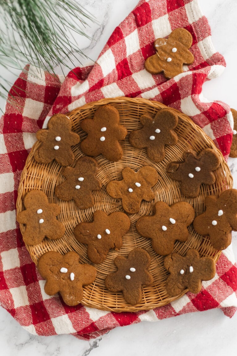 A round wicker tray filled with gingerbread cookies shaped like people, decorated with simple white icing dots, sits on a red and white checkered cloth on a white surface. Some pine needles are visible in the corner.