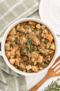 A white casserole dish filled with baked stuffing made of cubed bread and chopped celery, topped with a sprig of rosemary, sits on a green plaid cloth next to a white plate and wooden fork.