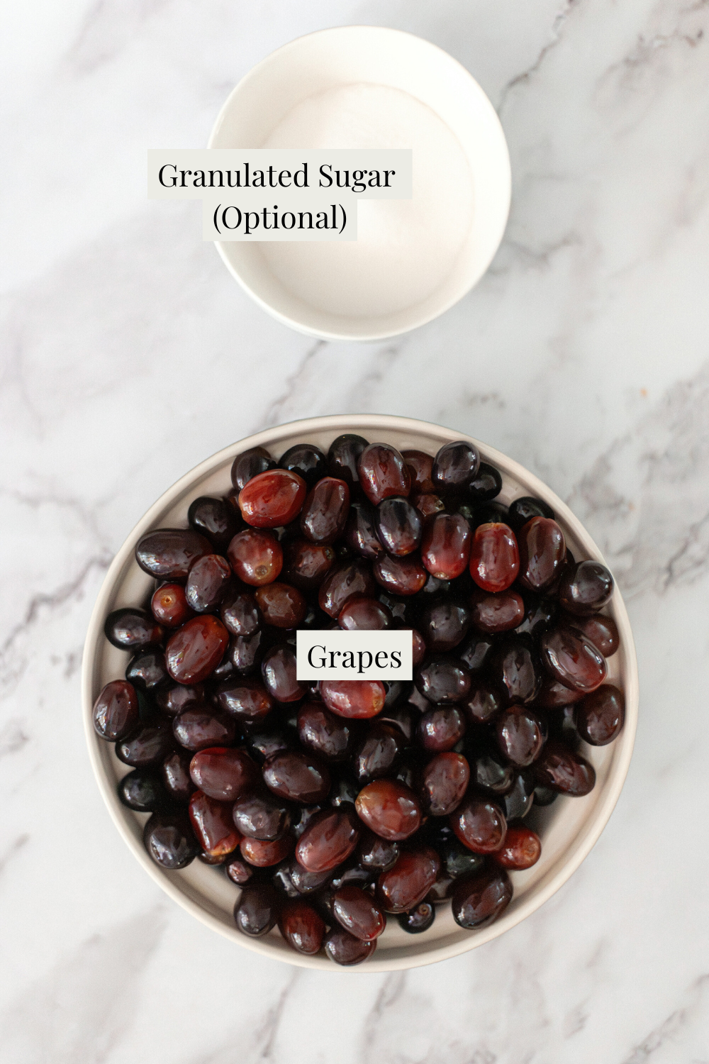 A bowl filled with dark grapes sits below a smaller bowl containing granulated sugar on a marble surface—perfect ingredients for learning How to Make and Can Grape Juice. Labels indicate Grapes and Granulated Sugar (Optional).