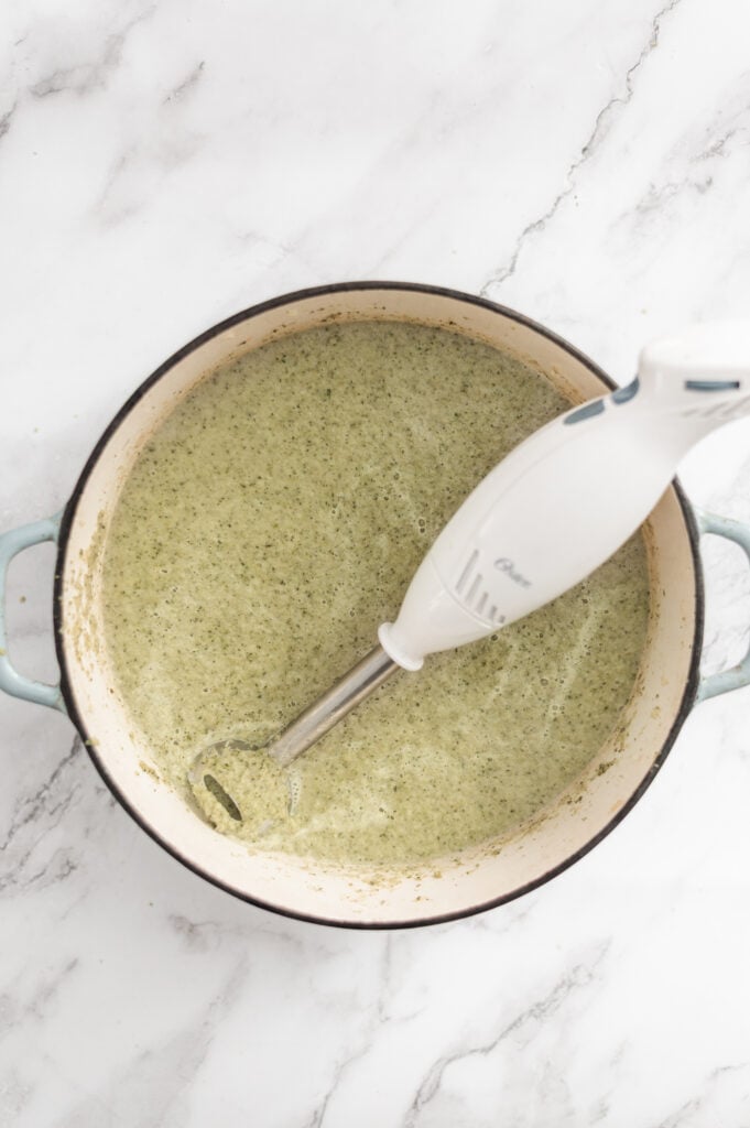 An immersion blender sits in a large pot filled with creamy green Cream of Broccoli Soup, placed on a white marble surface.
