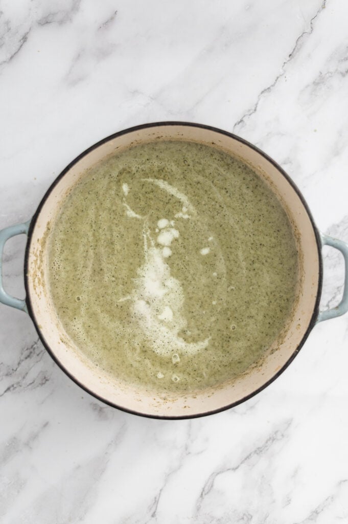 A large pot with blue handles contains Cream of Broccoli Soup, its creamy green surface swirled with lighter cream, shown from above on a white marble surface.