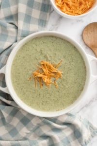 A bowl of creamy broccoli soup, made with rich Cream of Broccoli Soup and topped with shredded cheddar cheese, sits on a blue and white checkered cloth. A wooden spoon and a bowl of extra cheese rest nearby on the marble surface.