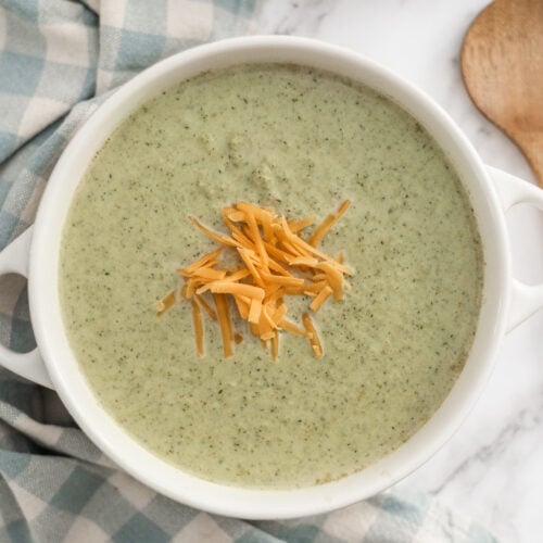 A bowl of creamy broccoli soup, made with rich Cream of Broccoli Soup and topped with shredded cheddar cheese, sits on a blue and white checkered cloth. A wooden spoon and a bowl of extra cheese rest nearby on the marble surface.