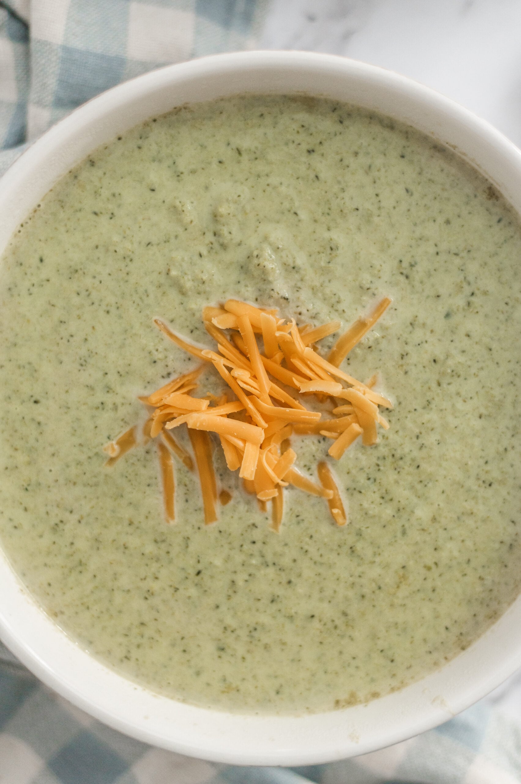 A bowl of classic Cream of Broccoli Soup topped with shredded cheddar cheese, placed on a blue and white checkered cloth.