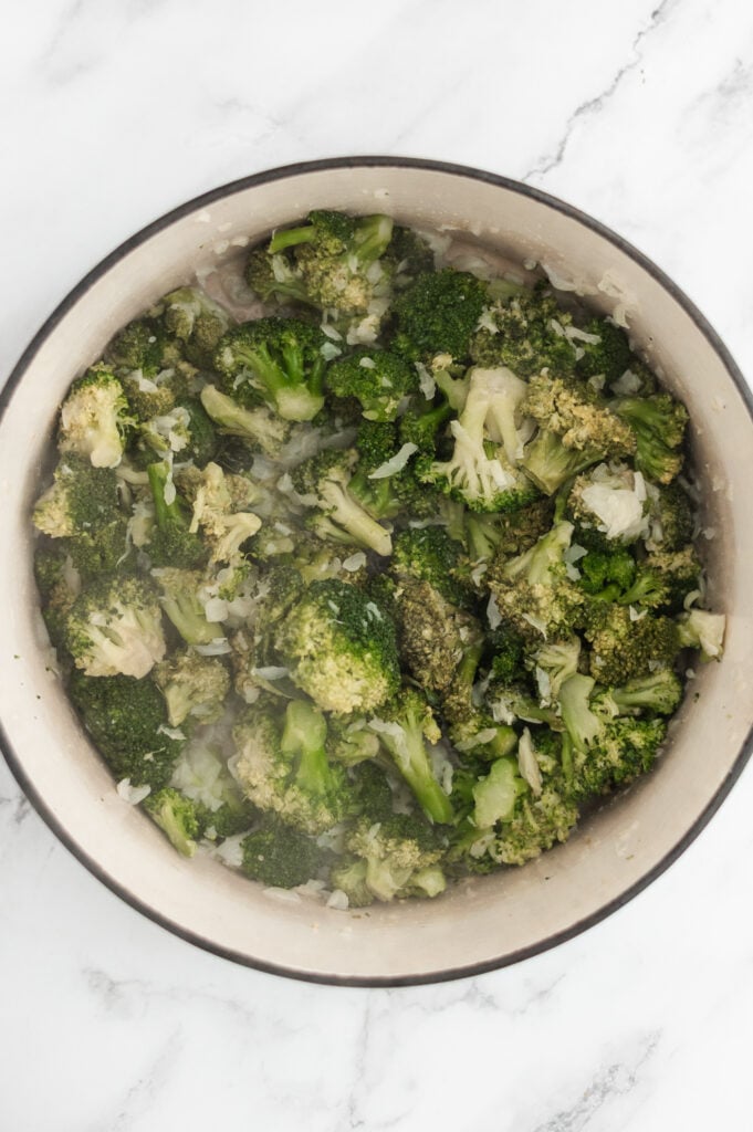 A bowl filled with cooked broccoli florets and small pieces of translucent onions, reminiscent of the fresh ingredients in Cream of Broccoli Soup, sits on a white marble surface.