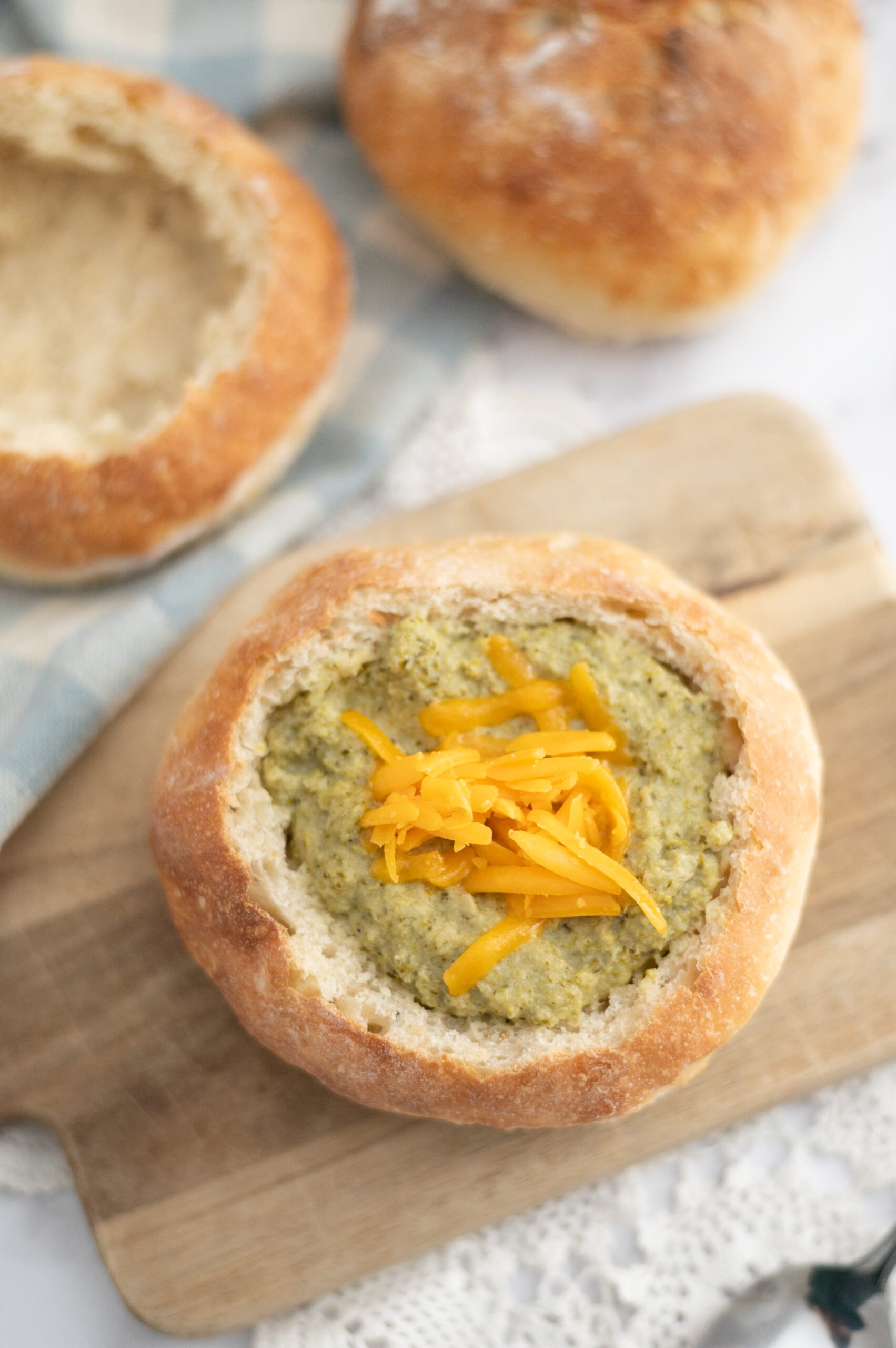 A bread bowl filled with creamy Cream of Broccoli Soup, topped with shredded cheddar cheese, sits on a wooden board. Another hollowed-out bread bowl is visible in the background, with a soft checkered cloth to the side.