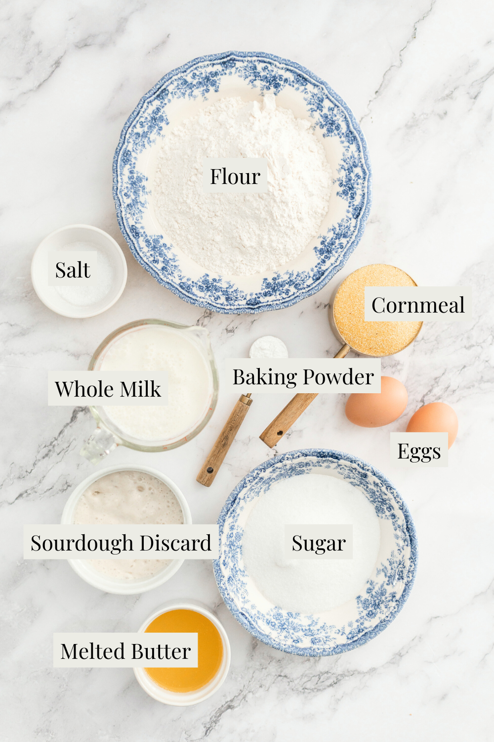A marble surface displays labeled bowls and containers with flour, cornmeal, sugar, salt, baking powder, whole milk, sourdough discard, melted butter, and eggs—everything neatly arranged to make delicious Sourdough Discard Cornbread.