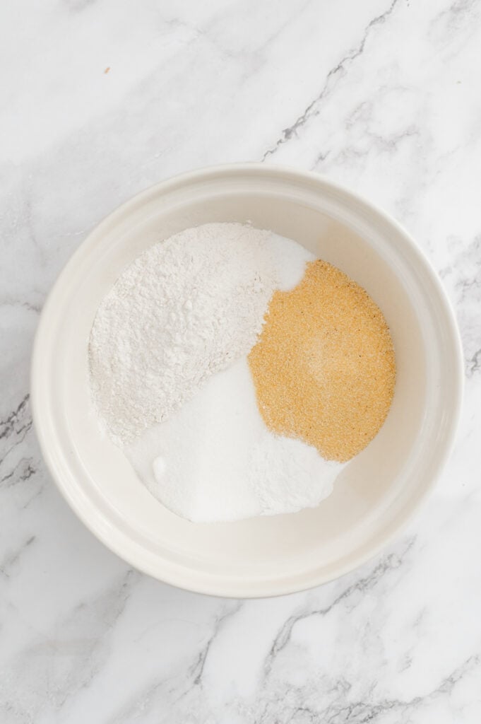 A white bowl on a marble surface contains three separate piles of dry ingredients for sourdough discard cornbread: flour, granulated sugar, and a yellow powder, possibly cornmeal.