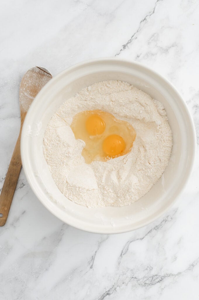 A white bowl with two raw eggs cracked into a well of flour for sourdough discard cornbread, next to a wooden spoon on a marble countertop.