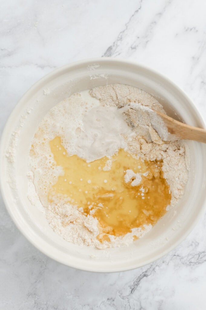 A mixing bowl with flour, oil, water, and sourdough discard being combined for cornbread with a wooden spoon on a marble surface.