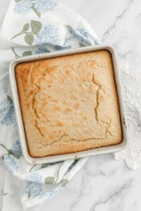 A golden-brown Sourdough Discard Cornbread sits in a metal baking pan on a white marble surface, with a blue floral napkin partially underneath. The cornbread's top is slightly cracked, hinting at its tender crumb beneath.