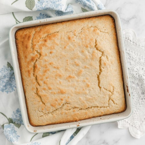 A golden-brown Sourdough Discard Cornbread sits in a metal baking pan on a white marble surface, with a blue floral napkin partially underneath. The cornbread's top is slightly cracked, hinting at its tender crumb beneath.