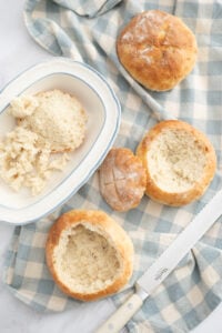 Three round Sourdough Discard Bread Bowls, two with tops cut off and insides hollowed out, rest on a blue and white checkered cloth. A knife and a plate with scooped-out bread pieces are also visible.