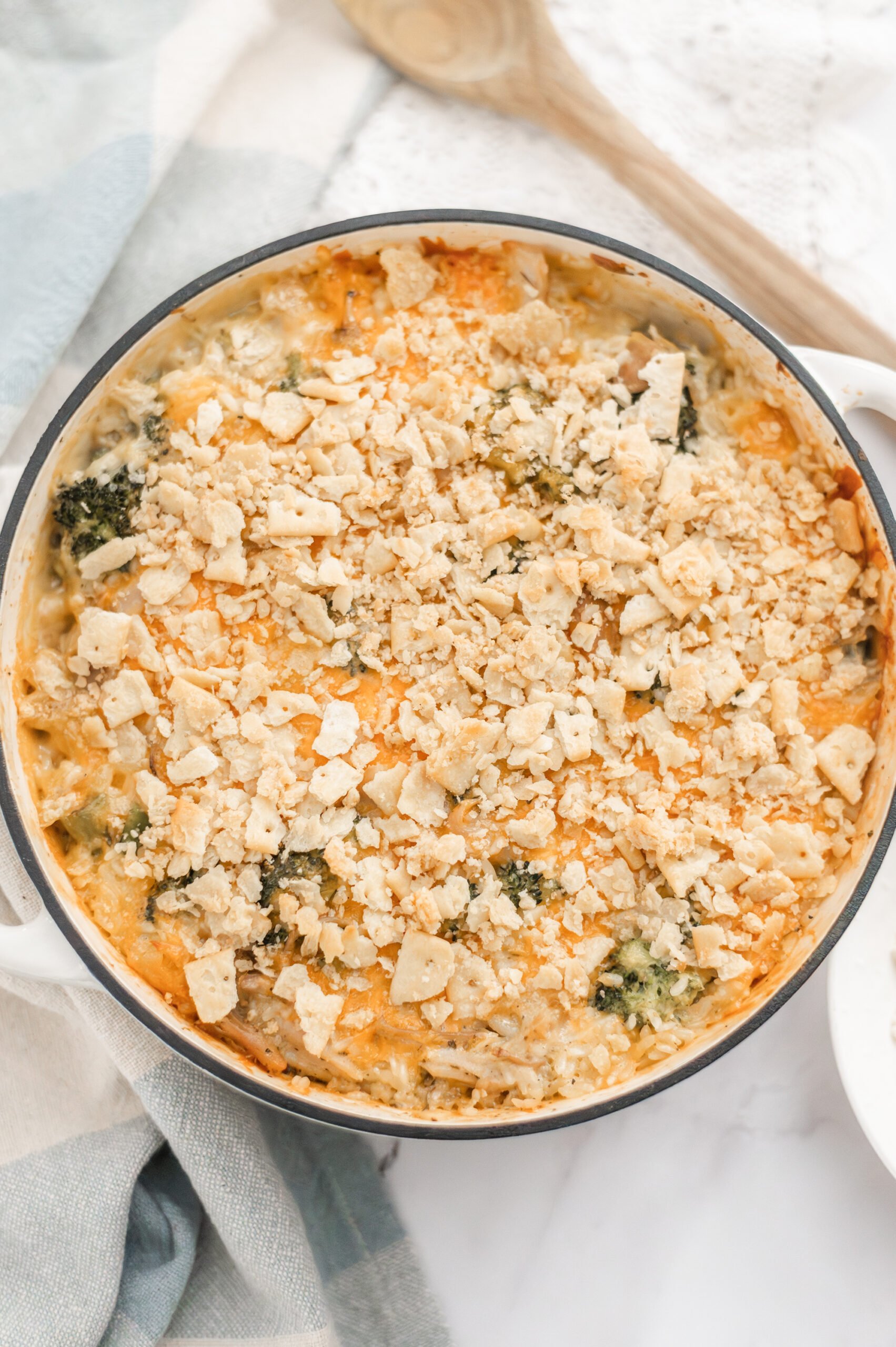 A baked Broccoli Chicken and Rice Casserole in a round white dish, topped with crushed crackers. The creamy casserole reveals hints of green vegetables beneath the topping. A wooden spoon and napkin are nearby.