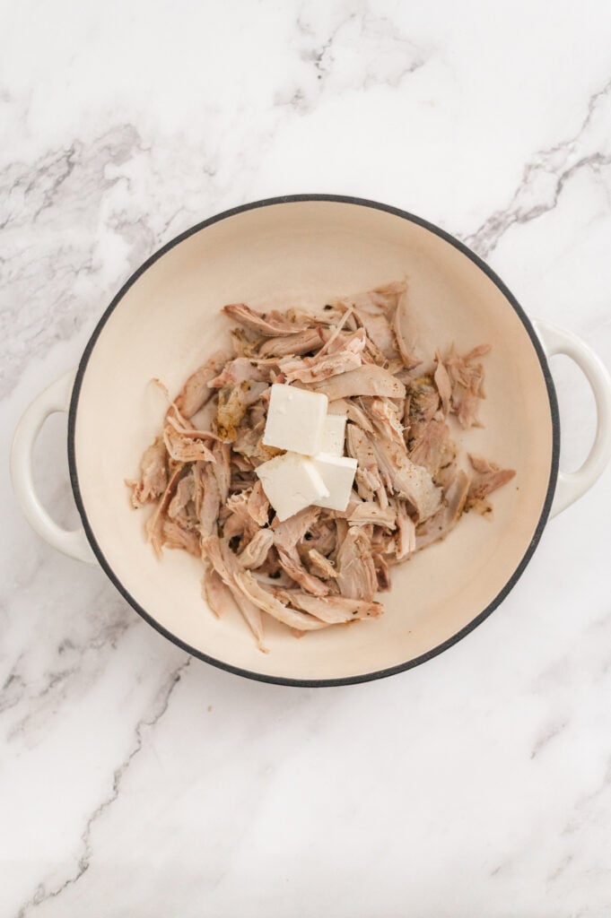 A white pot filled with shredded cooked meat and three cubes of cream cheese sits on a white marble surface, ready to be transformed into a delicious Broccoli Chicken and Rice Casserole.
