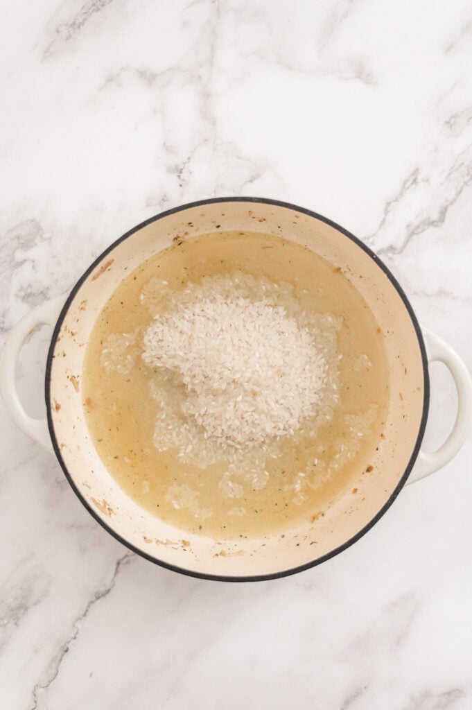 A white enameled pot with handles contains uncooked white rice and water on a white marble surface, ready to become the base for a delicious Broccoli Chicken and Rice Casserole.