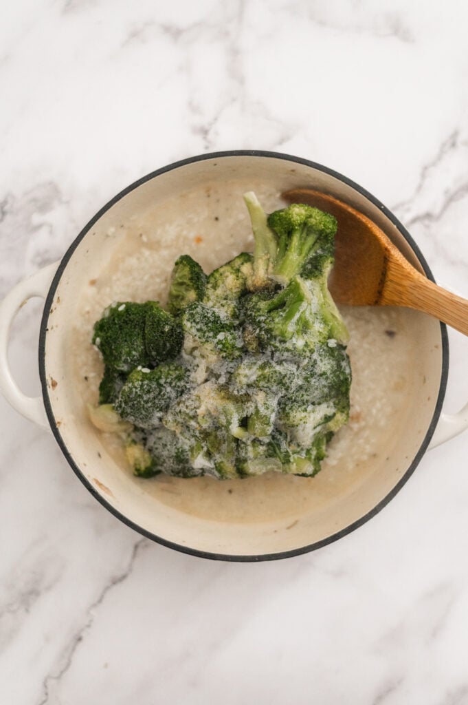 A white pot containing steamed broccoli topped with melted butter, reminiscent of the fresh flavors found in a Broccoli Chicken and Rice Casserole, with a wooden spoon resting on the side, set on a white marble surface.
