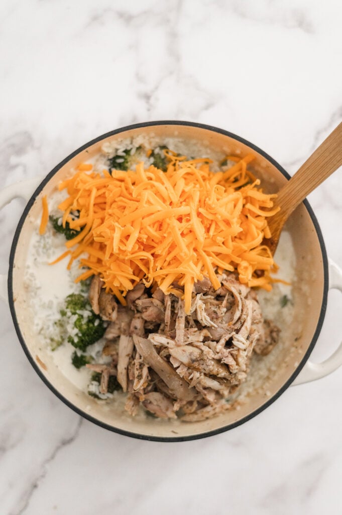 A pot filled with shredded cheddar cheese, cooked chicken, broccoli, creamy sauce, and rice is being mixed with a wooden spoon—perfect for making Broccoli Chicken and Rice Casserole on a white marble surface.